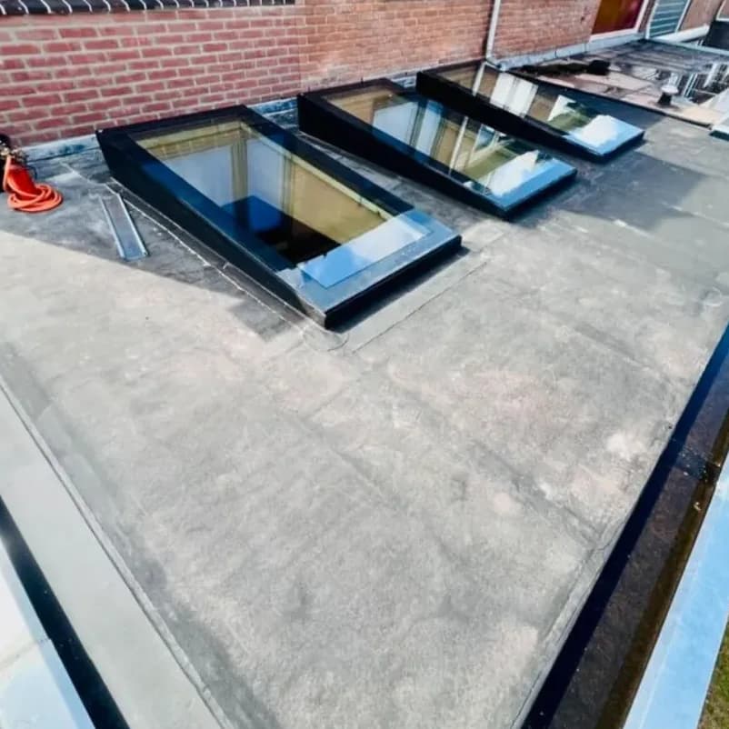 Plat bitumen dak met drie zwart omrande lichtkoepels langs een bakstenen gevel.