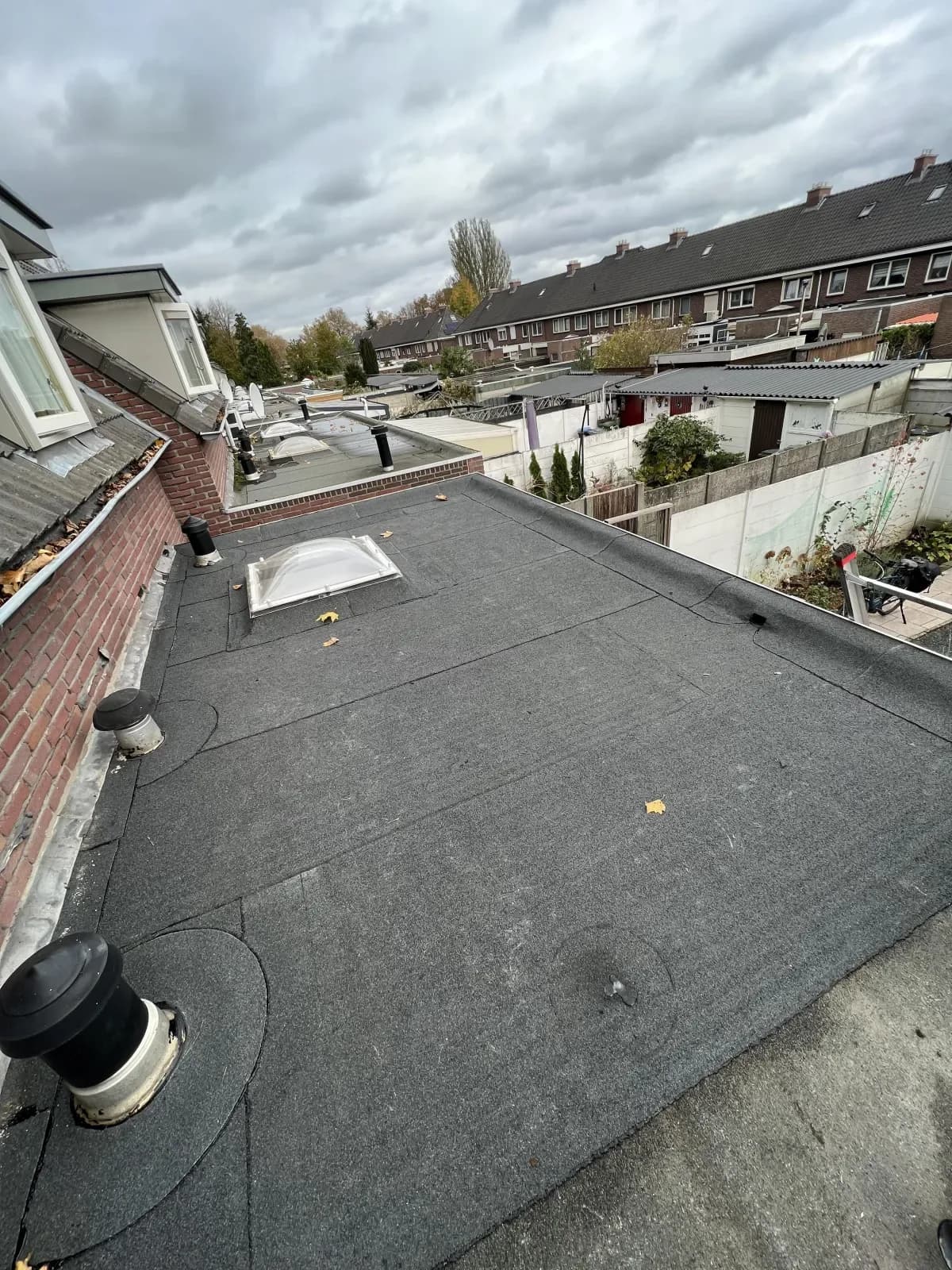 Nieuw aangebracht plat bitumen dak op een aanbouw, met lichtkoepel en ontluchtingspijpen, achter een rij woningen.